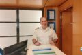 Smiling man in a white uniform with shoulder epaulettes behind a reception desk in a modern office.