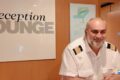Smiling hotel receptionist in a white uniform with shoulder epaulets at a front desk beside a large 'Reception' sign. (Informative)