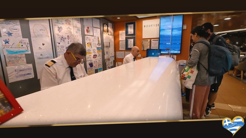 blue-star-ferries-tinos-aktoploikes-grammes-eisitiria 13 | Magic Lamp Reception desk at a travel lobby with two staff in white shirts assisting travelers with backpacks nearby; wall filled with children's drawings behind them