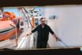 Man standing on a white ship deck beside an orange lifeboat, wearing black clothing and sunglasses, with railing on either side.