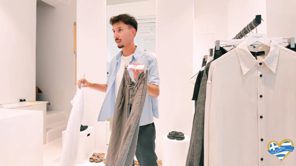 Man shopping in a bright clothing store, holding a white shirt and a pair of gray pants near a rack of clothes.