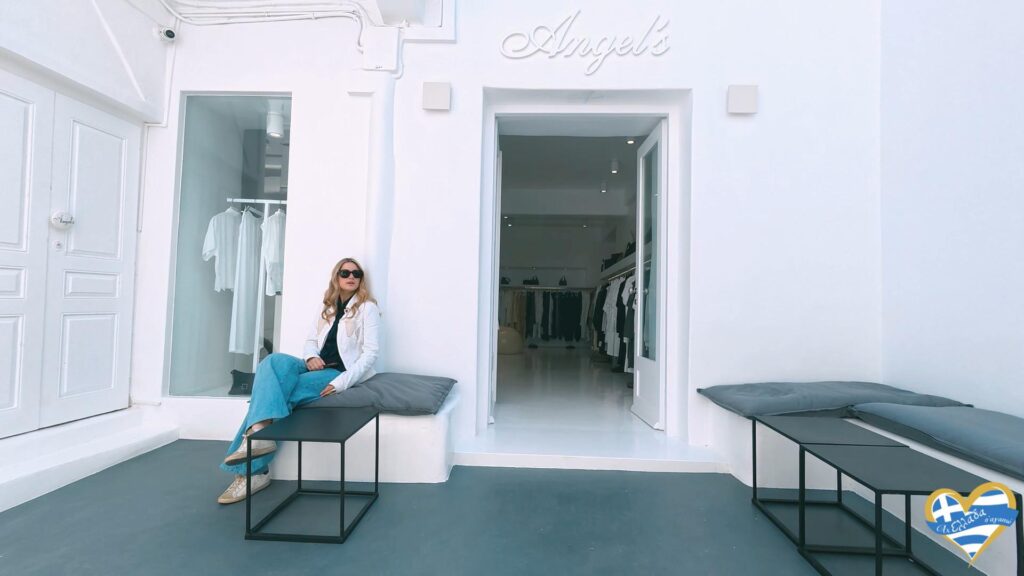 Woman in a white jacket and blue jeans sits on a bench outside a white clothing boutique with clothes visible through the open door.