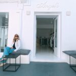 Woman in a white jacket and blue jeans sits on a bench outside a white clothing boutique with clothes visible through the open door.
