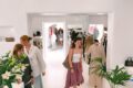 Shoppers browse clothes in a bright white boutique with handbag displays and plants in the foreground.