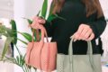 Person in a dark sweater holds two pastel handbags (pink and mint green) with woven handles in a bright shop.