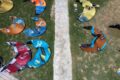 Colorful kites laid out on a grassy field around a central stone path, seen from above, likely preparing for a kiteboarding event; bottom right shows a heart-shaped blue-and-yellow logo.