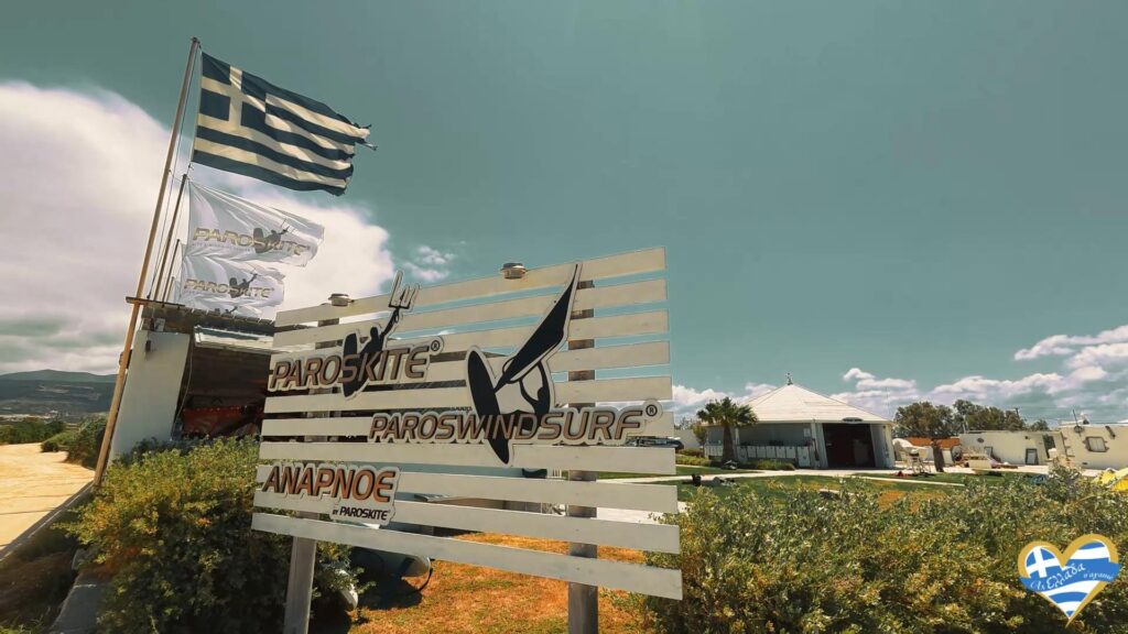 Signboard advertising Paroskite windsurfing/paros kite on a sunny day with flags nearby and a white building in the background