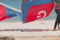 Person in a black wetsuit stands on a sandy beach with a red-blue windsurfing sail near the water. A heart-shaped logo is in the bottom right corner.