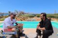 Two men sit and chat beside a bright turquoise pool on a sunny day, outdoor garden setting, with Paros Kite logos in the corner.