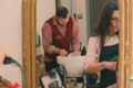 Barber in a burgundy vest washes a client’s hair at a salon sink, with a stylist in the foreground nearby.