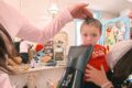 Child in a red shirt sits on a chair while a stylist trims his hair in a bright boutique salon, with two adults nearby watching or assisting.