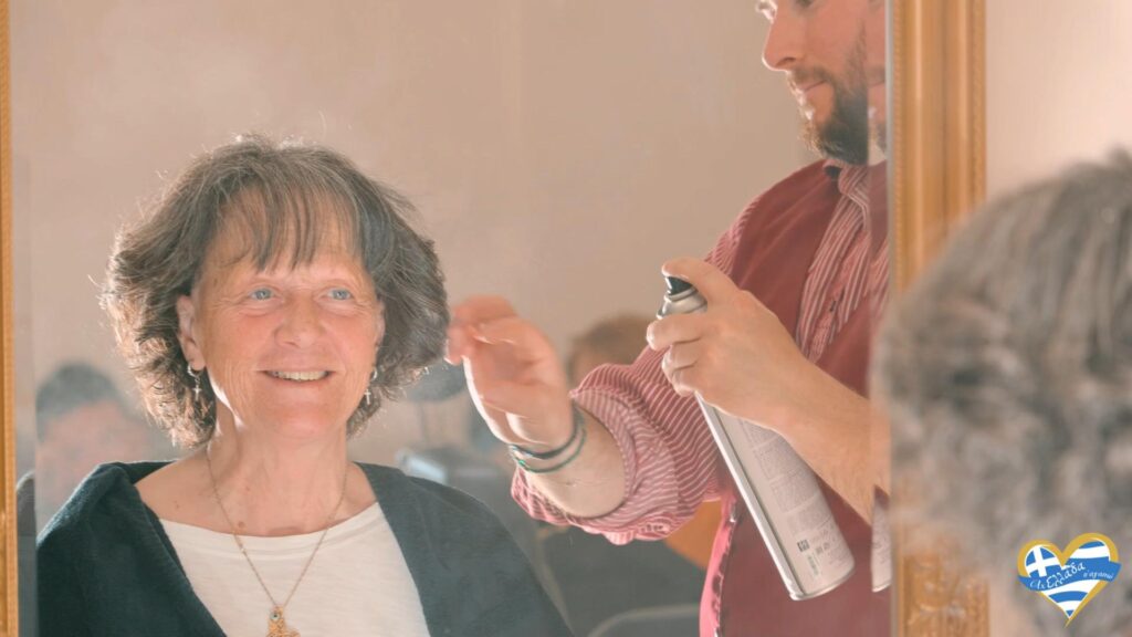 kommotirio-syros-paris-palais-de-beaute 5 | Magic Lamp Smiling older woman seated in a salon chair as a hairdresser sprays her hair with a can, seen in the mirror behind them.