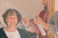 Smiling older woman seated in a salon chair as a hairdresser sprays her hair with a can, seen in the mirror behind them.