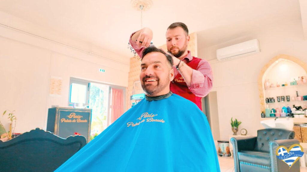 kommotirio-syros-paris-palais-de-beaute 6 | Magic Lamp Smiling man sits in a barber chair with a blue cape as a barber trims his hair indoors.