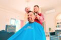 Smiling man sits in a barber chair with a blue cape as a barber trims his hair indoors.
