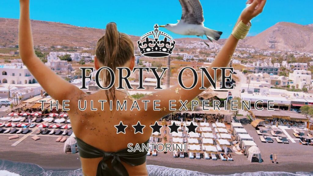 restaurant-beach-bar-santorini-forty-one 11 | Magic Lamp Back view of a woman with arms raised overlooking a Santorini beach club and white buildings, with promotional text'FORTY ONE' and 'THE ULTIMATE EXPERIENCE'.