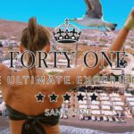 restaurant-beach-bar-santorini-forty-one 11 | Magic Lamp Back view of a woman with arms raised overlooking a Santorini beach club and white buildings, with promotional text 'FORTY ONE' and 'THE ULTIMATE EXPERIENCE'.