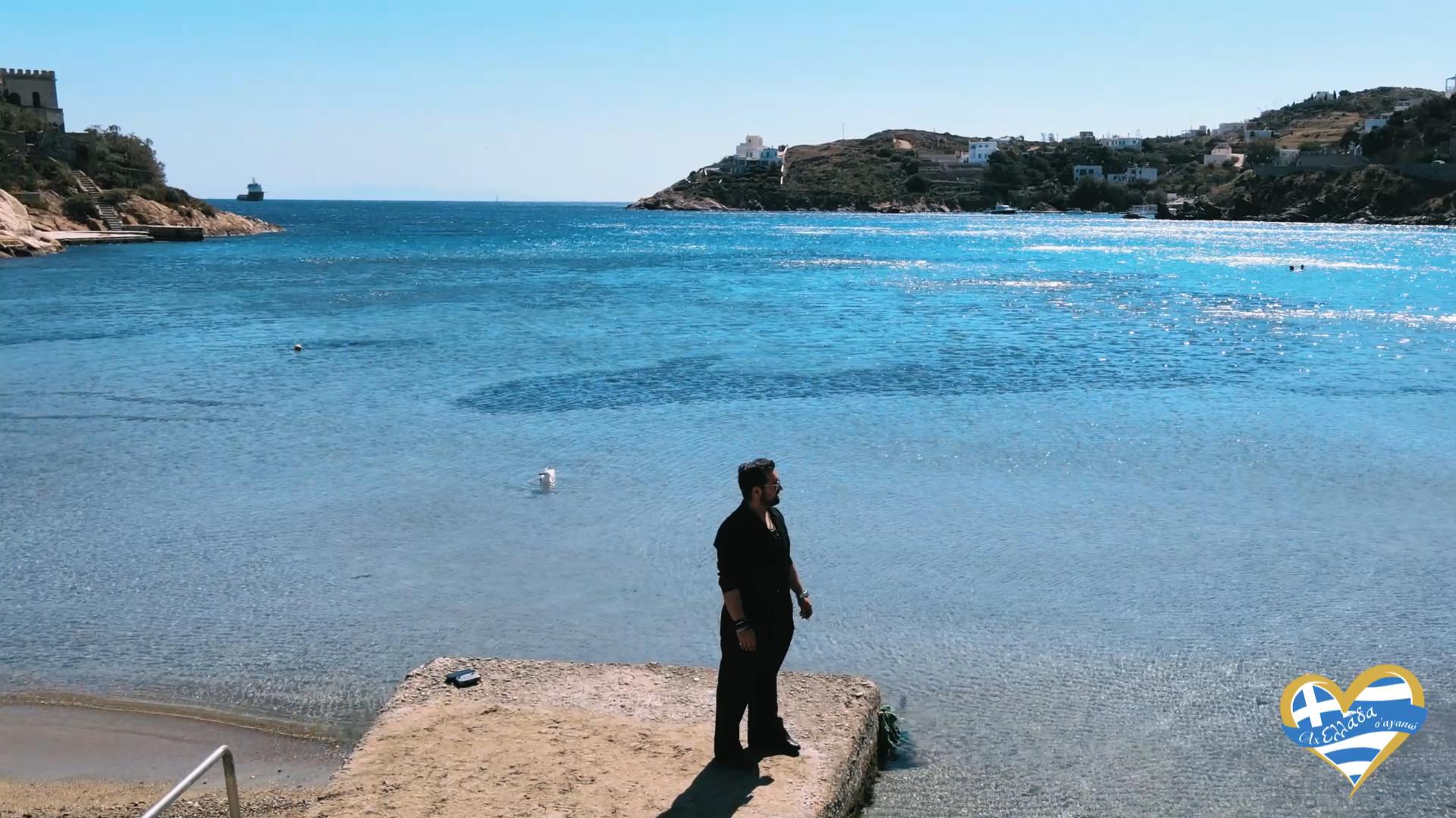 Man in dark clothing stands on a concrete pier by clear, turquoise sea with a coastal town in the background, sunny sky above.