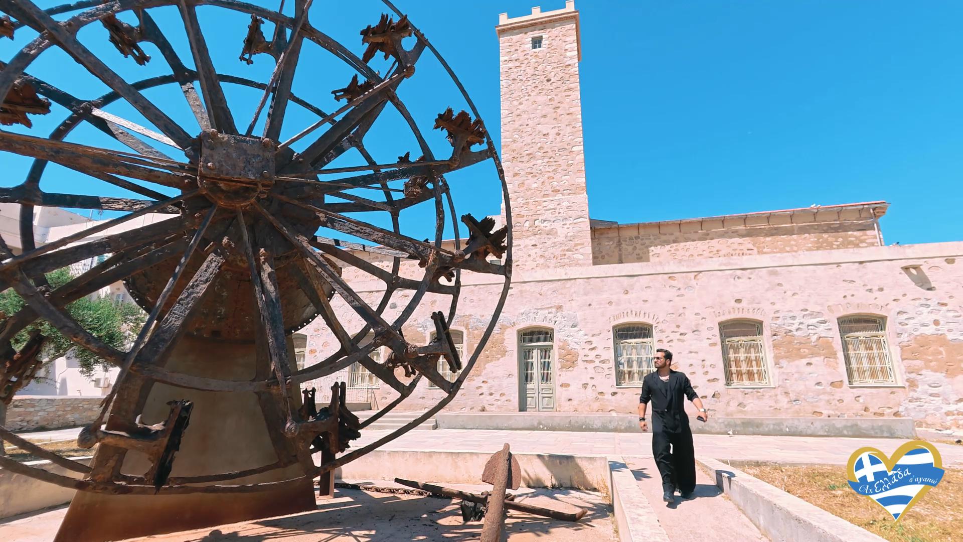 Rusty iron spherical wheel sculpture in a courtyard beside a beige stone building with blue sky; a man in black stands nearby.