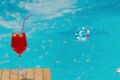 Red cocktail in a stemmed glass with a purple straw, placed on a wooden surface beside a bright blue pool behind. The 'Summer Shades' logo is visible in the background.