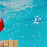 Red cocktail in a stemmed glass with a purple straw, placed on a wooden surface beside a bright blue pool behind. The 'Summer Shades' logo is visible in the background.