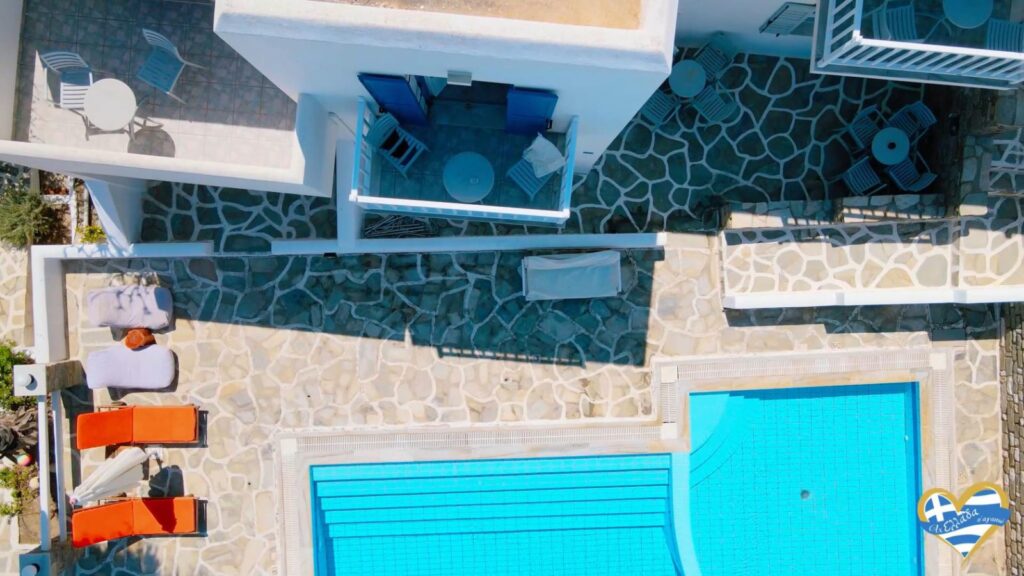 Aerial view of a white-walled resort courtyard with a bright blue rectangular pool, stone-paved decks, orange lounge chairs, and white tables/chairs on upper terraces.