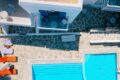 Aerial view of a white-walled resort courtyard with a bright blue rectangular pool, stone-paved decks, orange lounge chairs, and white tables/chairs on upper terraces.