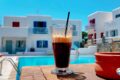 Glass of iced coffee with a black straw on a wooden poolside table, blue pool and white modern buildings in the background.