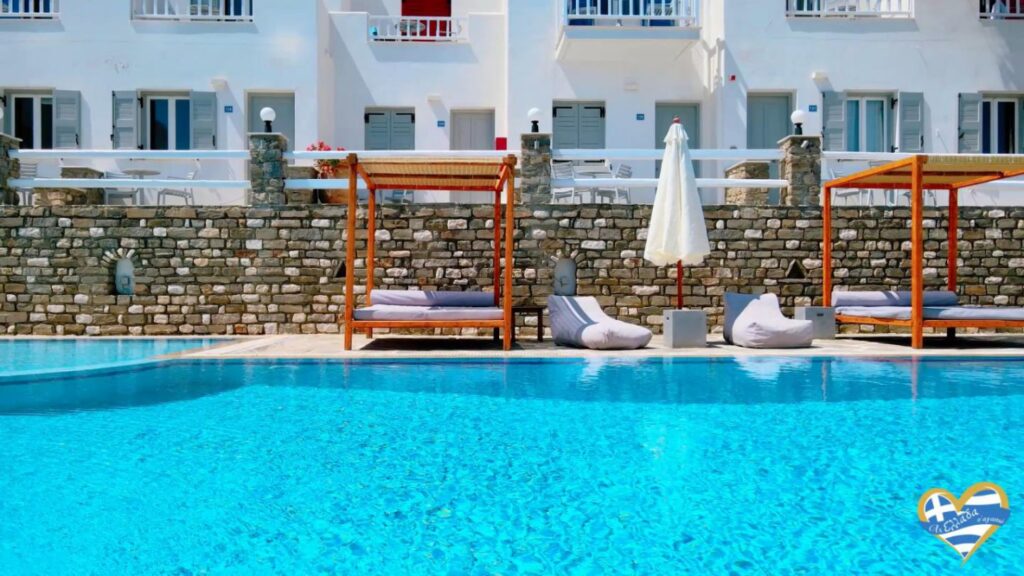 Crystal blue swimming pool with wooden cabanas and a stone wall backdrop, white buildings, and lounge seating along the pool deck.