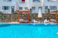 Crystal blue swimming pool with wooden cabanas and a stone wall backdrop, white buildings, and lounge seating along the pool deck.