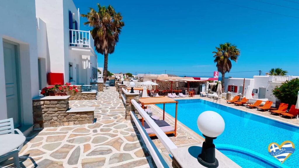 White-washed hotel courtyard with a bright blue pool, stone walkways, and orange loungers under clear skies.