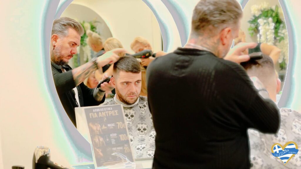 Barber trims a man's hair at a salon station, reflected in a large circular mirror with other clients visible in the background.