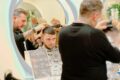 Barber trims a man's hair at a salon station, reflected in a large circular mirror with other clients visible in the background.
