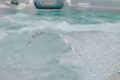 Close-up of a large clear ice chunk resting on a spa pool, with frothy water in the background and a circular logo in the bottom-right corner.