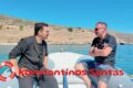 Two men sit on a small boat near a rocky coastline, wearing sunglasses, with clear blue water and sky; red text overlay reads 'Konstantinos Santas'. A life ring is on the left and a logo appears bottom-right.