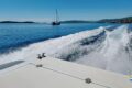 View from a speeding boat with a wake in the foreground, a sailboat ahead on calm blue water, and a coastline in the distance.