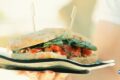 Close-up of a rustic sandwich filled with greens and tomato, held on a glossy plate with two wooden skewers piercing the bread.