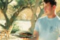 Man wearing a light blue shirt holds a plate with a tall, stacked burger outdoors, with wooden chairs and trees in the background.