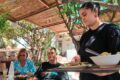 Waitress in black serves a plate of salad to two guests at an outdoor cafe under a wooden pergola.