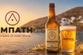 Beer advertisement banner featuring a bottle and glass of Amphati beer on a rocky shoreline at sunset with splashing water and a coastal town in the background