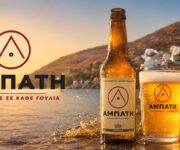 Beer advertisement banner featuring a bottle and glass of Amphati beer on a rocky shoreline at sunset with splashing water and a coastal town in the background