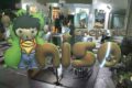 Nighttime storefront with a giant green cartoon boy statue and oversized metallic letters spelling the shop name, with customers visible inside.