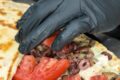 Gloved hand placing chopped tomatoes and olives into a flatbread wrap with meat filling.