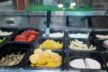Self-serve deli counter with multiple trays of food, including corn, sliced tomatoes, fried chicken, and mushrooms, with a serving knife in the center.