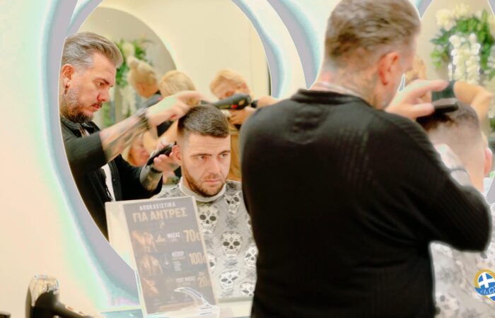 Barber trims a man's hair at a salon station, reflected in a large circular mirror with other clients visible in the background.
