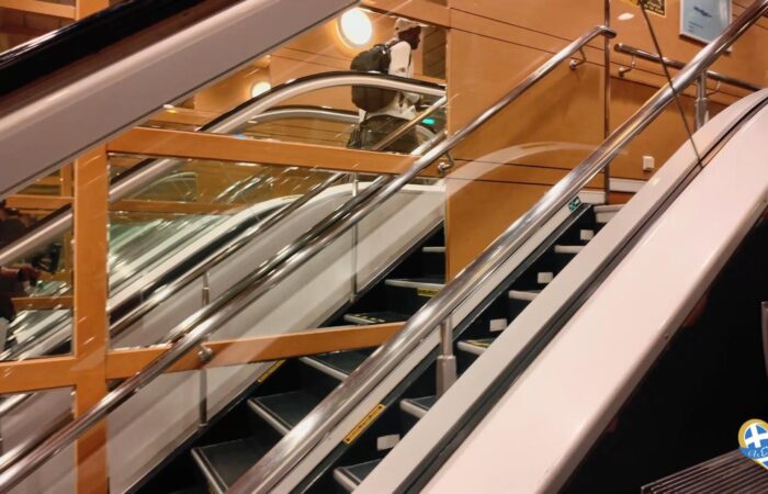 Indoor multi-level atrium with wooden railings and chrome escalators; a person wearing a backpack stands on the upper escalator