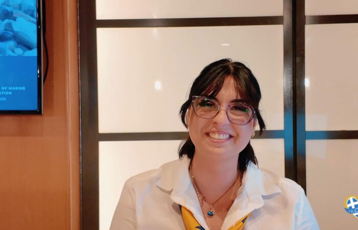 Smiling woman with dark hair and glasses, wearing a white shirt and yellow scarf, posed indoors.