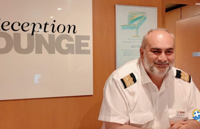 Smiling hotel receptionist in a white uniform with shoulder epaulets at a front desk beside a large 'Reception' sign. (Informative)