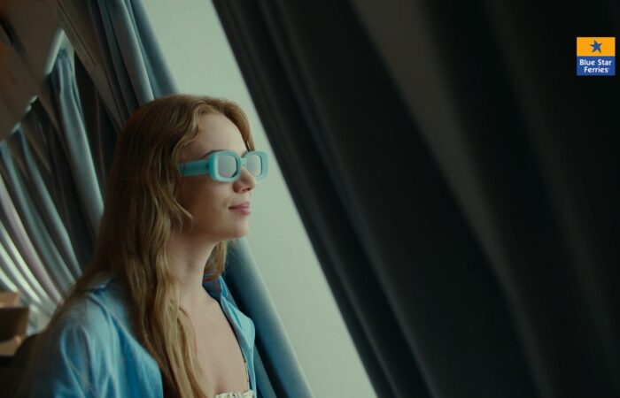 Woman wearing turquoise 3D-style glasses gazing out the ferry window, Blue Star Ferries logo in the top-right corner