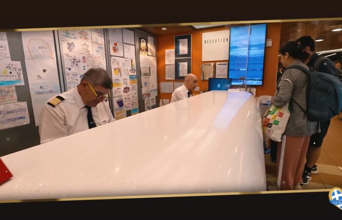 Reception desk at a travel lobby with two staff in white shirts assisting travelers with backpacks nearby; wall filled with children's drawings behind them